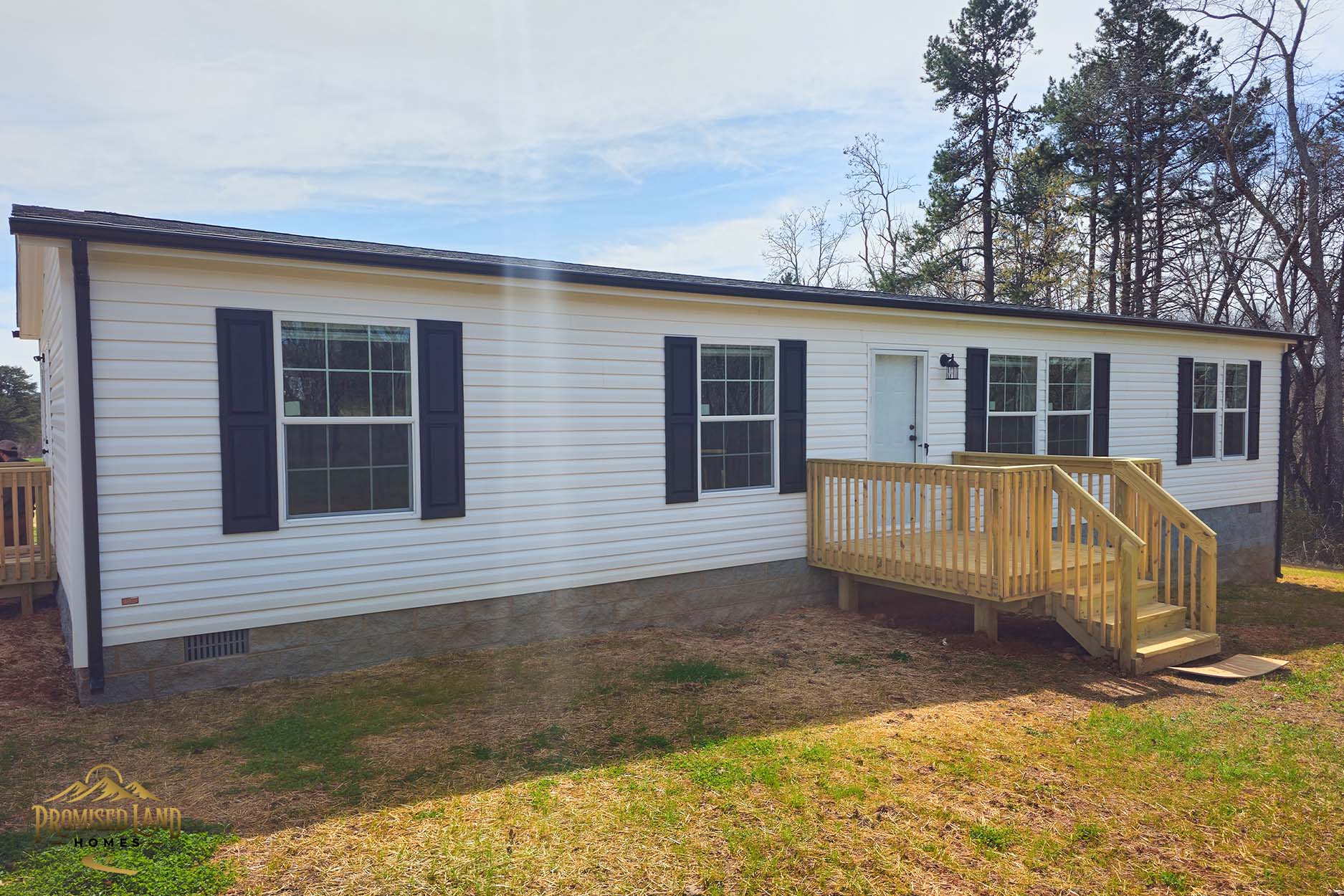 A white manufactured home with black shutters and a wooden ramp entrance sits on a grassy lot surrounded by trees.